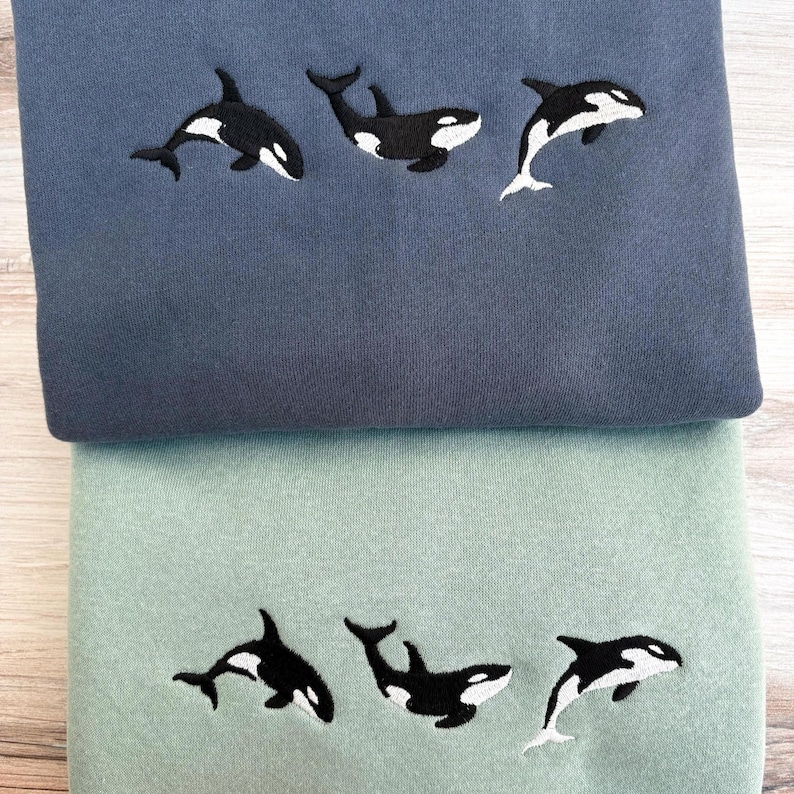 May include: Two sweatshirts, one blue and one green, each embroidered with three black and white orca whale designs. The whales are in different poses, with some leaping and others swimming. The sweatshirts are folded and stacked.