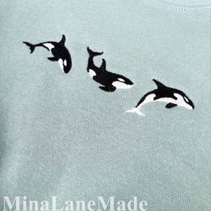 May include: Pale blue t-shirt with three embroidered orcas in black and white. The orcas are arranged in a swimming pattern. The text "MinaLaneMade" is at the bottom of the image.