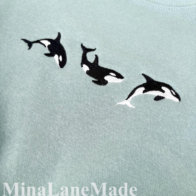 May include: Pale blue t-shirt with three embroidered orcas in black and white. The orcas are arranged in a swimming pattern. The text "MinaLaneMade" is at the bottom of the image.