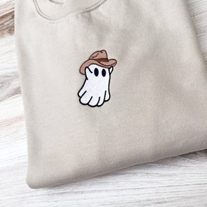 May include: A beige sweatshirt with a small embroidered patch of a white ghost wearing a brown cowboy hat. The ghost has two black eyes and a black outline. The sweatshirt is folded neatly on a light-colored wooden surface.