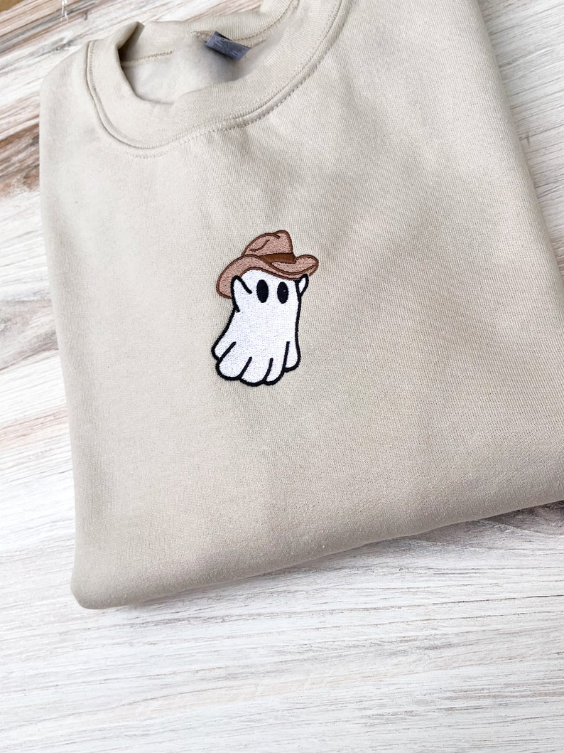 May include: A beige sweatshirt with a small embroidered patch of a white ghost wearing a brown cowboy hat. The ghost has two black eyes and a black outline. The sweatshirt is folded neatly on a light-colored wooden surface.