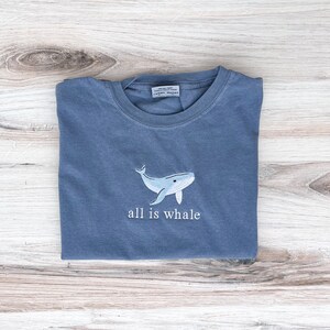 Może przedstawiać: Niebieska koszulka z jasnoniebieskim haftem przedstawiającym wieloryba i napisem "all is whale". Koszulka jest złożona na jasnej drewnianej powierzchni. Projekt jest marynistyczny.