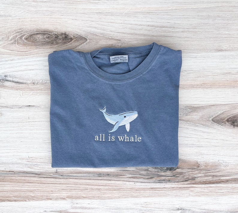 Może przedstawiać: Niebieska koszulka z jasnoniebieskim haftem przedstawiającym wieloryba i napisem "all is whale". Koszulka jest złożona na jasnej drewnianej powierzchni. Projekt jest marynistyczny.
