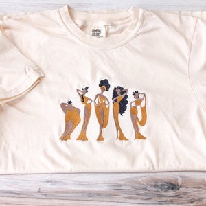 May include: A cream colored t-shirt with an embroidered design of five women in gold dresses. The text "COMFORT COLORS" is on the tag.