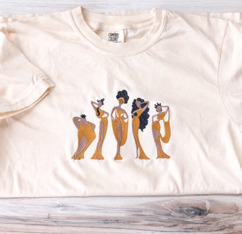 May include: A cream colored t-shirt with an embroidered design of five women in gold dresses. The text "COMFORT COLORS" is on the tag.