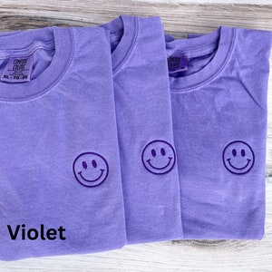 May include: Three purple t-shirts with a smiling face embroidered on the front. The t-shirts are folded on a wooden surface. The text "Violet" is written below the shirts.