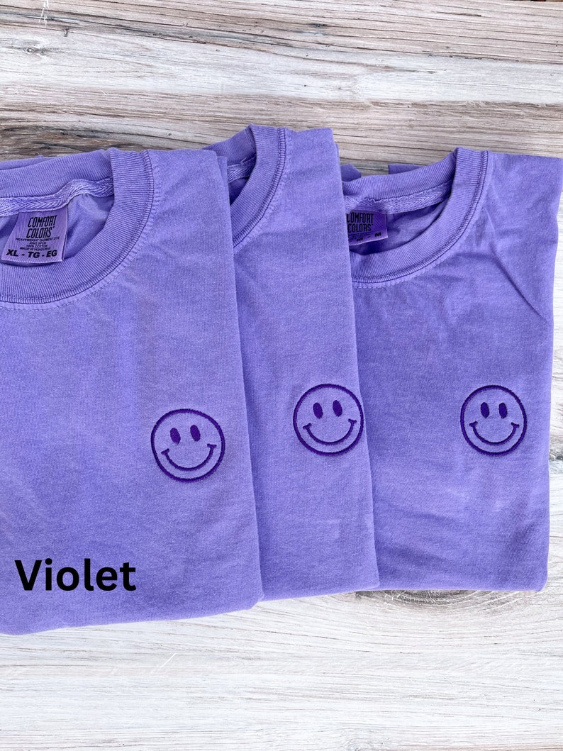 May include: Three purple t-shirts with a smiling face embroidered on the front. The t-shirts are folded on a wooden surface. The text "Violet" is written below the shirts.