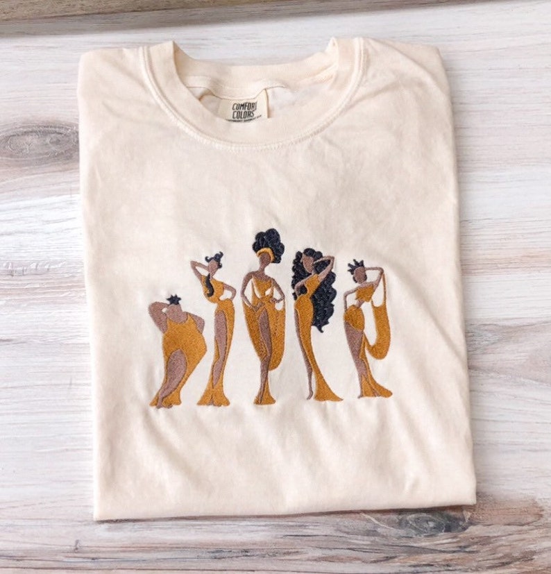 May include: A cream colored t-shirt with an embroidered design of five women in gold dresses. The women are standing in a line and have dark skin and black hair. The text "COMFORT COLORS" is visible on the tag.