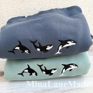 May include: Two folded sweatshirts, one blue and one green, stacked. Each sweatshirt features three embroidered black and white orca whales. The bottom of the image has the text "MinaLaneMade".