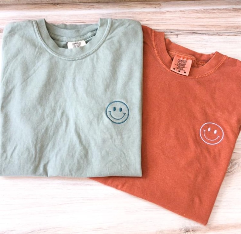 May include: Two t-shirts with a smiley face embroidered on the front. The shirt on the left is a light green color and the shirt on the right is a burnt orange color.