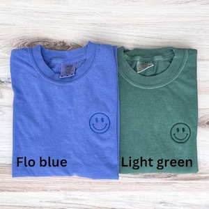 May include: Two t-shirts, one light blue and one light green, with a smiling face embroidered on the front. The light blue shirt has the text "Flo blue" printed below the smiley face, and the light green shirt has the text "Light green" printed below the smiley face.