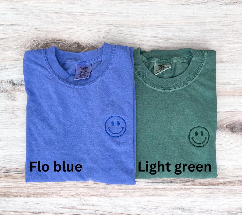 May include: Two t-shirts, one light blue and one light green, with a smiling face embroidered on the front. The light blue shirt has the text "Flo blue" printed below the smiley face, and the light green shirt has the text "Light green" printed below the smiley face.