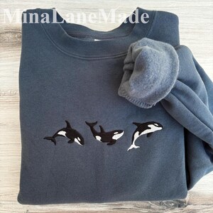 May include: A slate blue sweatshirt featuring three embroidered orca whales in black and white. The text "MinaLaneMade" is visible at the top. The sweatshirt is folded, with the sleeve bunched up.