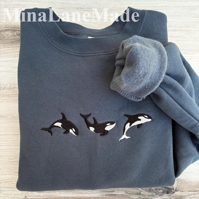 May include: A slate blue sweatshirt featuring three embroidered orca whales in black and white. The text "MinaLaneMade" is visible at the top. The sweatshirt is folded, with the sleeve bunched up.