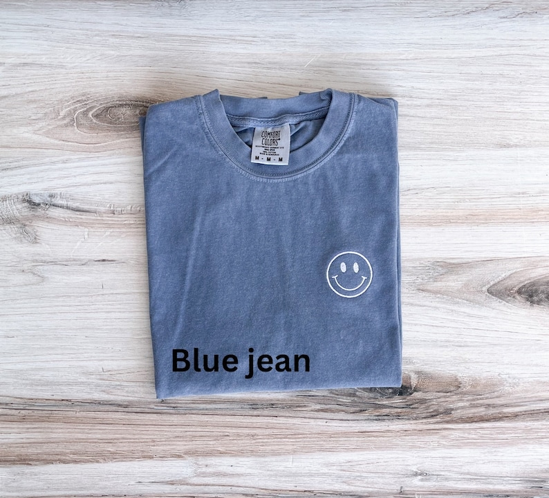 May include: A blue jean t-shirt with a white smiley face embroidered on the front. The shirt is folded and laid flat on a wooden surface.