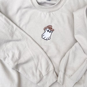 May include: A beige crewneck sweatshirt featuring an embroidered ghost wearing a brown cowboy hat. The ghost has two black eyes and a black outline. The sweatshirt has long sleeves and a ribbed neckline, cuffs, and hem.