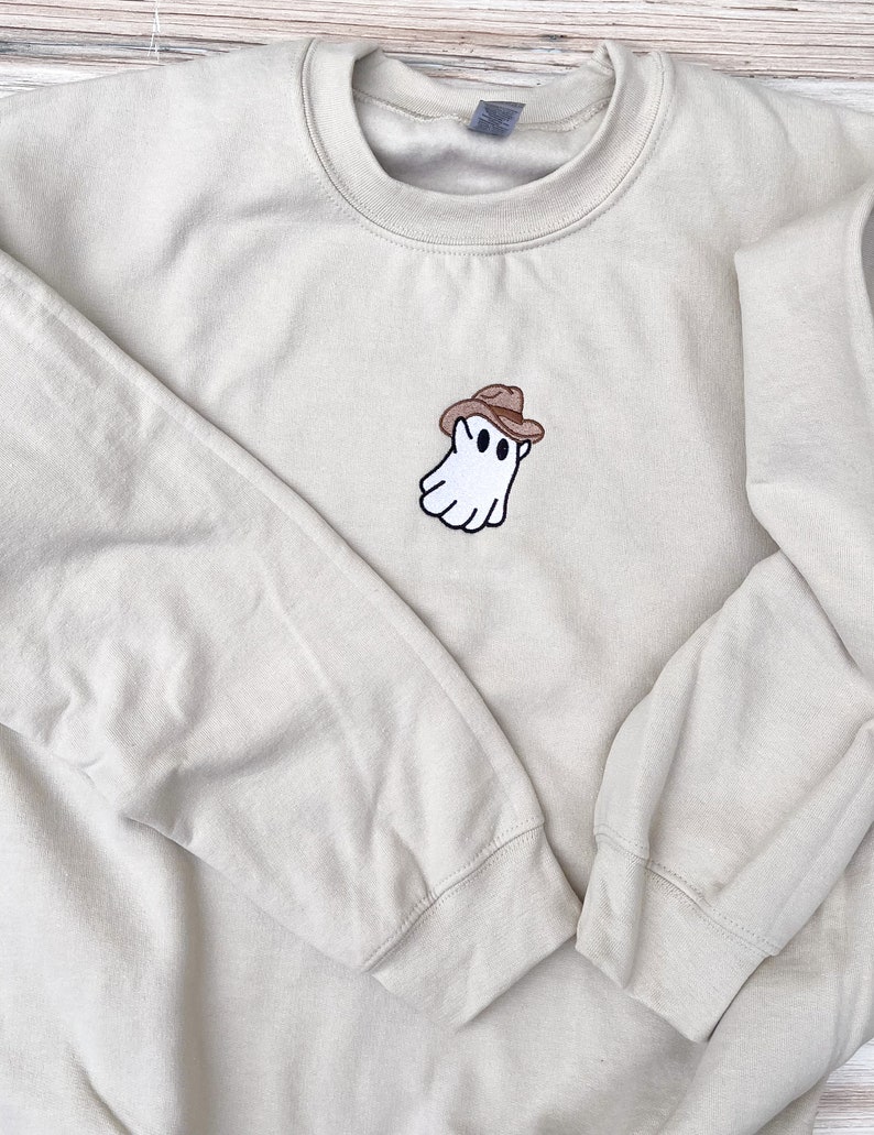 May include: A beige crewneck sweatshirt featuring an embroidered ghost wearing a brown cowboy hat. The ghost has two black eyes and a black outline. The sweatshirt has long sleeves and a ribbed neckline, cuffs, and hem.