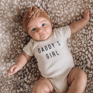 May include: A baby wearing a beige bodysuit with the text "Daddy's Girl" in black. The baby is wearing a brown turban-style headband and is lying on a brown and white patterned blanket.