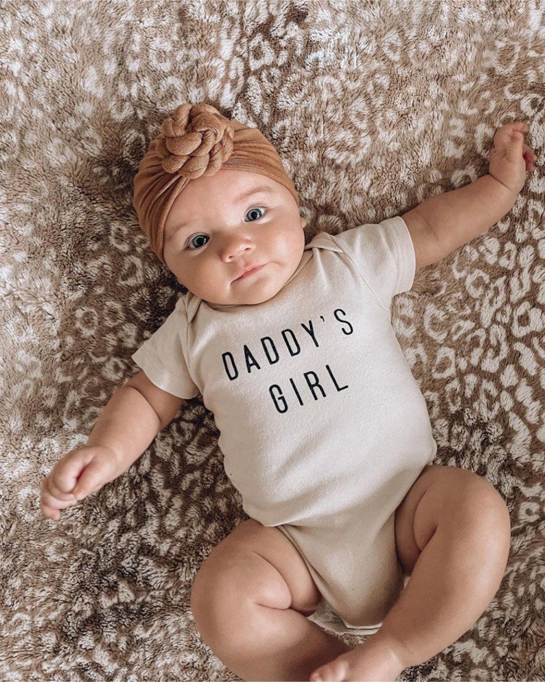 May include: A baby wearing a beige bodysuit with the text "Daddy's Girl" in black. The baby is wearing a brown turban-style headband and is lying on a brown and white patterned blanket.