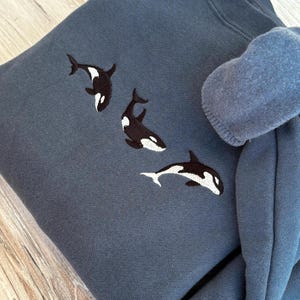 May include: A dark blue sweatshirt with three embroidered orca whales in black and white. The whales are displayed across the front. The sleeve is folded over, revealing the inside lining.