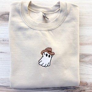 May include: A beige sweatshirt with a small embroidered design of a white ghost wearing a brown cowboy hat. The ghost has two black eyes and a smiling mouth. The sweatshirt is folded neatly.