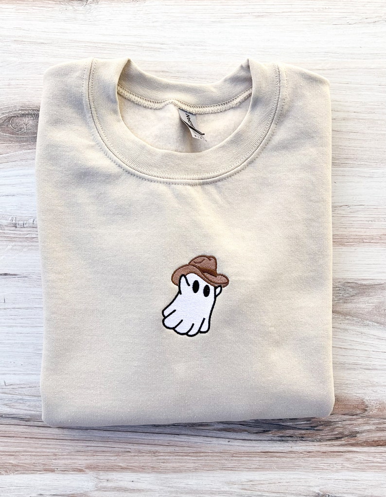 May include: A beige sweatshirt with a small embroidered design of a white ghost wearing a brown cowboy hat. The ghost has two black eyes and a smiling mouth. The sweatshirt is folded neatly.