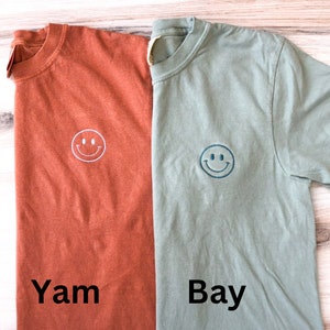 May include: Two t-shirts with a smiley face embroidered on the front. The shirt on the left is rust colored and says "Yam" below the smiley face. The shirt on the right is sage green and says "Bay" below the smiley face.