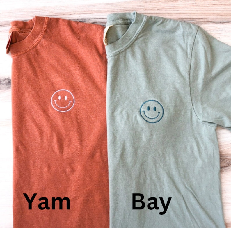 May include: Two t-shirts with a smiley face embroidered on the front. The shirt on the left is rust colored and says "Yam" below the smiley face. The shirt on the right is sage green and says "Bay" below the smiley face.