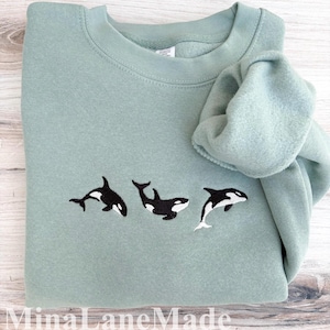 May include: A sage green sweatshirt with an embroidered design of three black and white orcas. The orcas are in different poses, with one leaping out of the water. The sweatshirt is folded, with the sleeve draped over the top.