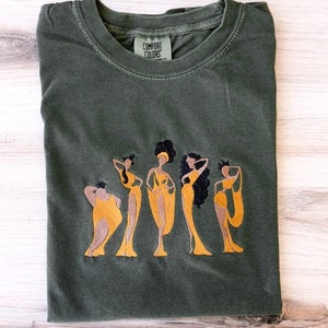 May include: A dark green t-shirt with an embroidered design of five women in yellow dresses. The text "COMFORT COLORS" is visible on the shirt.
