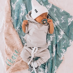 May include: A baby wearing a grey hoodie and white beanie sleeps on a green and white wave patterned blanket. The blanket has a pink fuzzy blanket underneath it with the word "mama" embroidered on it.