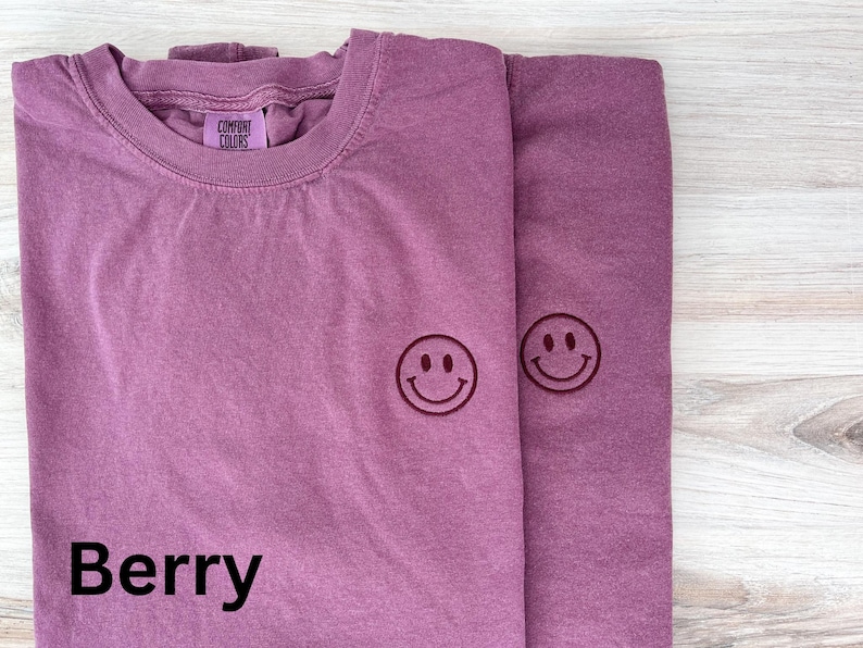 May include: Two purple t-shirts with a smiling face embroidered on each. The t-shirts are folded and the text "Berry" is printed on the bottom of the left t-shirt.