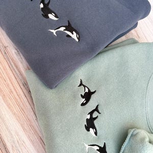 May include: Two sweatshirts, one in a dusty blue and the other in a soft green, are folded on a wooden surface. Each sweatshirt features an embroidered design of three black and white orca whales swimming downwards. The sweatshirts are made of a soft material.