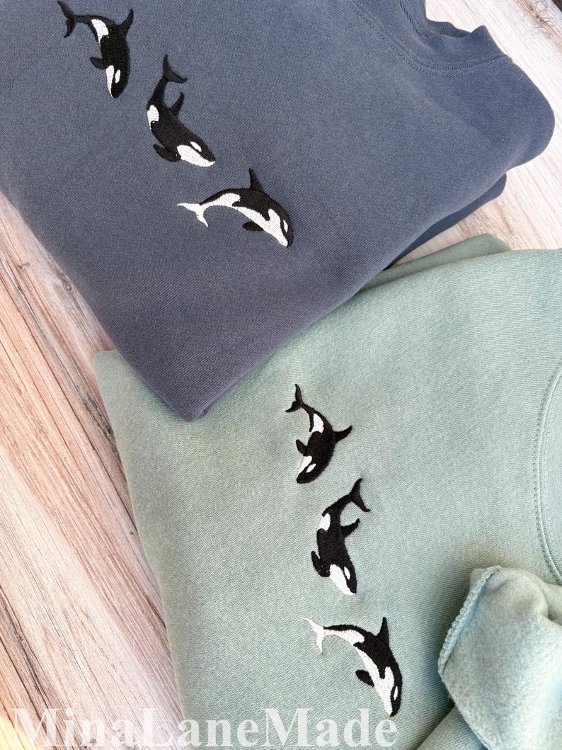 May include: Two sweatshirts, one in a dusty blue and the other in a soft green, are folded on a wooden surface. Each sweatshirt features an embroidered design of three black and white orca whales swimming downwards. The sweatshirts are made of a soft material.