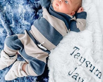 May include: A baby wearing a long-sleeve waffle knit shirt and pants with horizontal navy blue and beige stripes, paired with cream-colored booties. The baby lies on a plush blanket that is navy blue on one side and white with the name Teysum Jay embroidered on the other.