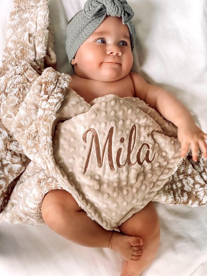 May include: A baby girl wearing a gray headband with a bow is wrapped in a beige and white plush blanket. The blanket has a brown embroidered name "Mila" on it.