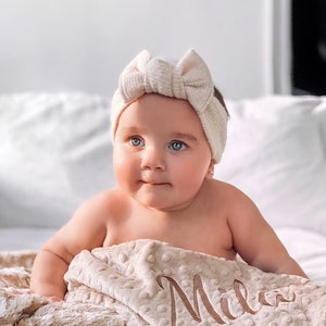 May include: A baby wearing a cream-colored headband with a bow sits on a cream-colored blanket with the name "Mila" embroidered on it.