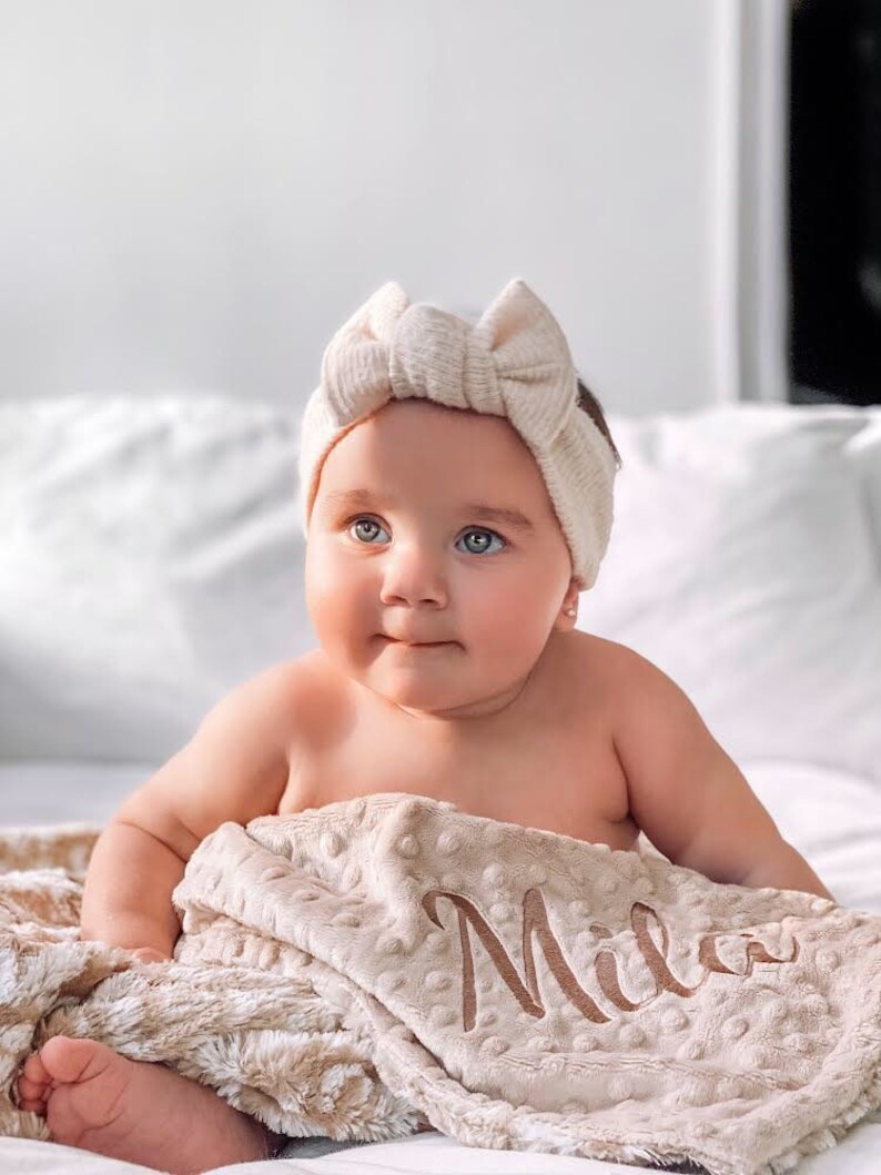 May include: A baby wearing a cream-colored headband with a bow sits on a cream-colored blanket with the name "Mila" embroidered on it.