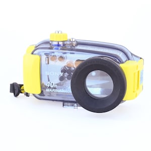 May include: A clear and yellow Sony Marine Pack underwater camera housing. The housing is designed for a Cyber-shot camera and is rated to 40m. The housing has a clear front lens and a black rubber ring around the lens.
