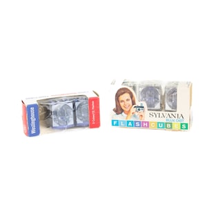 May include: Two boxes of flash cubes, one from Westinghouse and one from Sylvania Blue Dot. Both boxes feature a woman holding a camera and the text "Flash Cubes".
