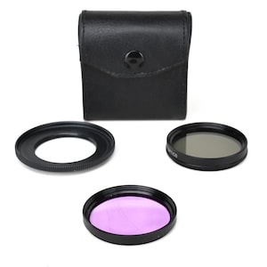 May include: A set of camera filters with a black leather case. The filters include a black ring, a gray filter, and a purple filter. The case is rectangular with a snap closure.
