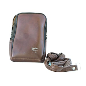 May include: A brown leather Kodak pouch case with a zip closure and a matching brown strap. The case has the words "Kodak POUCH CASE" printed on the front. The strap is looped and has a metal clasp.