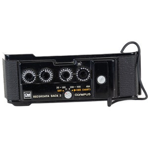 Puede incluir: Cámara Olympus negra con una perilla plateada y una correa negra. La cámara tiene una serie de diales con números y el texto "Recordata Back 2" y "Olympus".