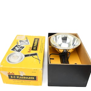May include: A vintage Kodak B-C Flashholder set, featuring a bright chrome reflector and a black handle. The flash unit is housed in a black box, while the yellow box displays the product name and a diagram.