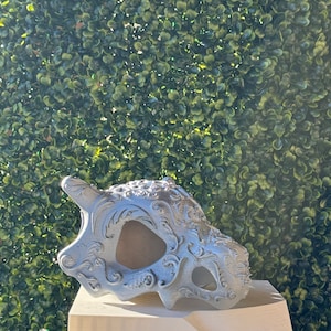 May include: A silver-coloured, detailed, 3D printed skull with intricate floral designs. The skull is sitting on a white platform.