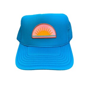 May include: A bright blue trucker hat with a pink, orange, and yellow sunset embroidered patch on the front.