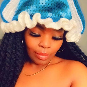 May include: A blue and white crocheted hat with a ruffled brim. The hat has a unique design with two white ear-like flaps.