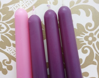 May include: Four taper candles in shades of pink and purple. The candles are arranged diagonally, with the pink candle on the left and the darkest purple on the right. The background is a white surface with a gold floral pattern.