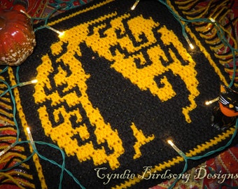 May include: A black and yellow crocheted square with a design of two cats. The square has a fringe border and is decorated with string lights.