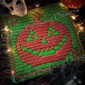 Puede incluir: Cuadrado de crochet verde y naranja con un diseño de calabaza pixelado. El cuadrado tiene un borde de flecos y el texto "Cynthie Birdsong Designs" es visible en la imagen.